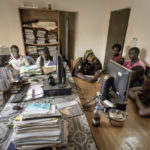 Im Büro der Hilfsorganisation "Association Malienne des Expulses" sind der AME-Mitarbeiter Amadou Coulibaly (1. Person v. links) und die ehrenamtliche AME-Mitarbeiterin Mariam Teme (4. Person v. links) mit der Registrierung der 6 Migranten beschäftigt.